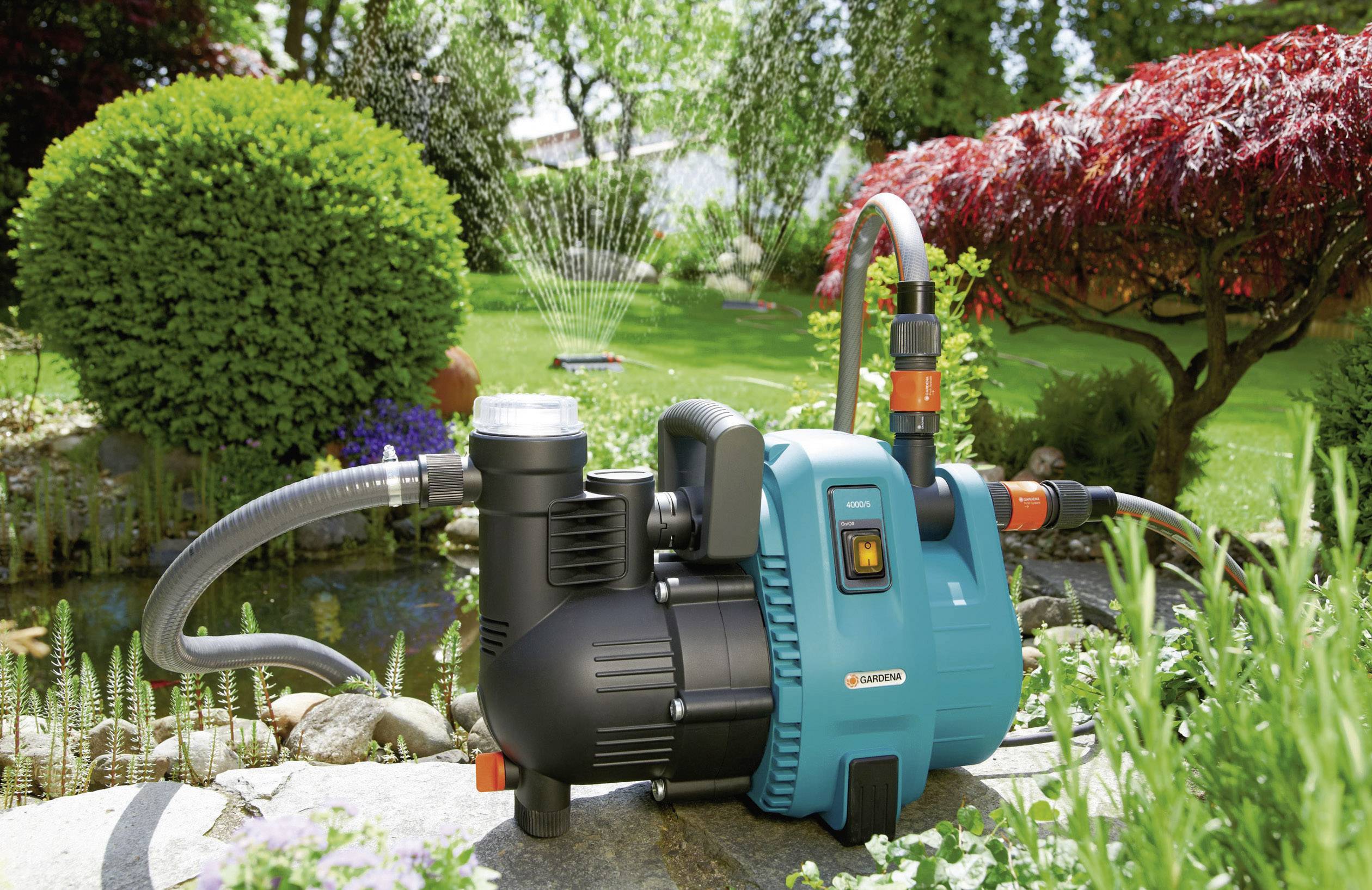 Zahradní zavlažovací čerpadlo před rybníkem, s připojenou hadicí, která zavlažuje trávník v slunečné, zelené zahradě.