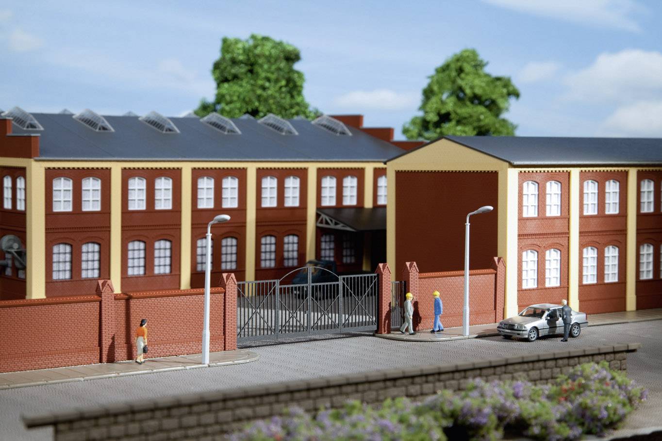 Model budovy továrny s červenou cihlovou fasádou a okny. Před vstupní bránou stojí lidé a automobily.