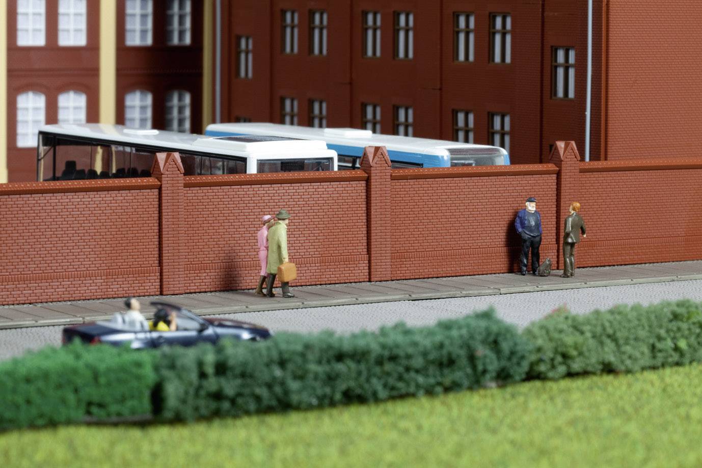 Miniaturní scéna se dvěma osobami stojícími u cihlové zdi; autobus a budovy v pozadí, automobil v popředí.