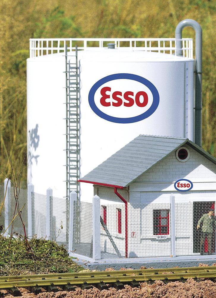 Velký bílý tankovací silo společnosti Esso s výrazným logem stojí v popředí před malou strážní budkou. V pozadí jsou trávy.