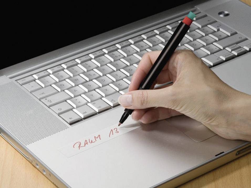 Ruka píše propiskou 'MÍSTNOST 13' na lepicí pásku nalepené na notebooku.