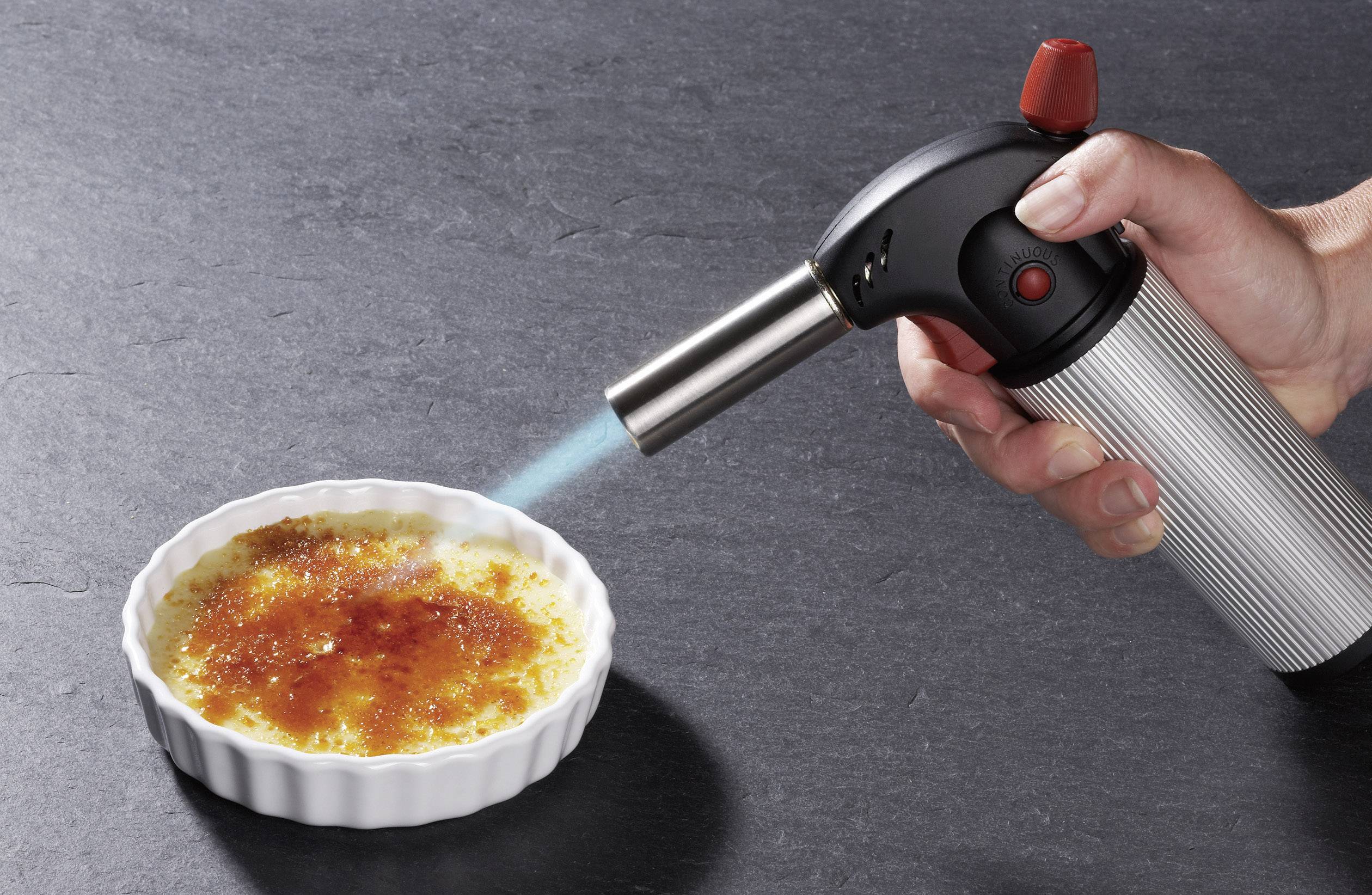 Ruka drží plynovou hořákem nad crème brûlée v bílé misce, zatímco se povrch karamelizuje.