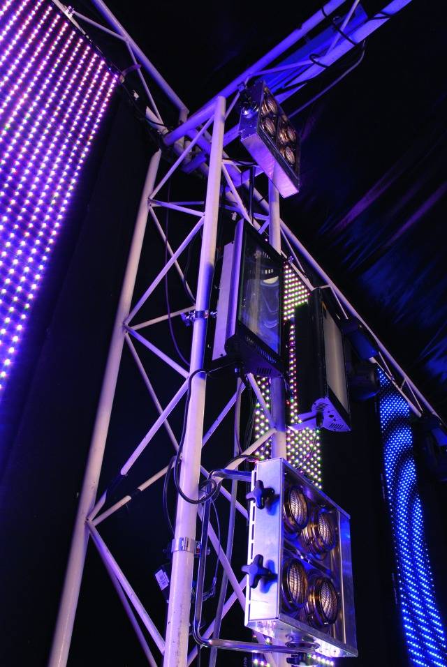 Světelná instalace s divadelními reflektory na kovovém lešení, osvětlená v modro-fialových tónech.