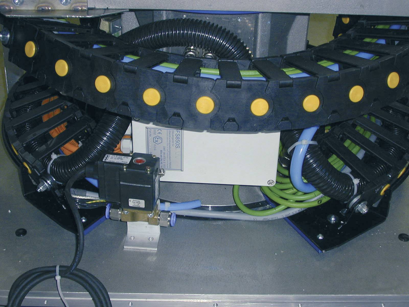 Mechanická součástka s kabely a hadicemi. Nahoře jsou viditelné černé nosníky se žlutými body. Níže se nachází elektronický modul.