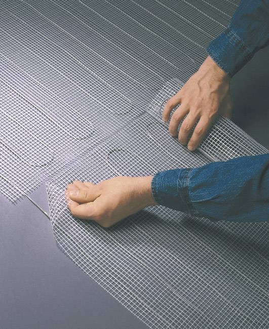 Osoba rozvaluje mřížovou síť na šedé podlaze, pravděpodobně v souvislosti se stavebními pracemi nebo pokládkou podlahy.