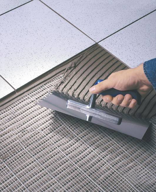 Ruka nanáší pomocí lžíce lepidlo na síť pro obkládačky. Vlevo nahoře jsou již položené bílé dlaždice.