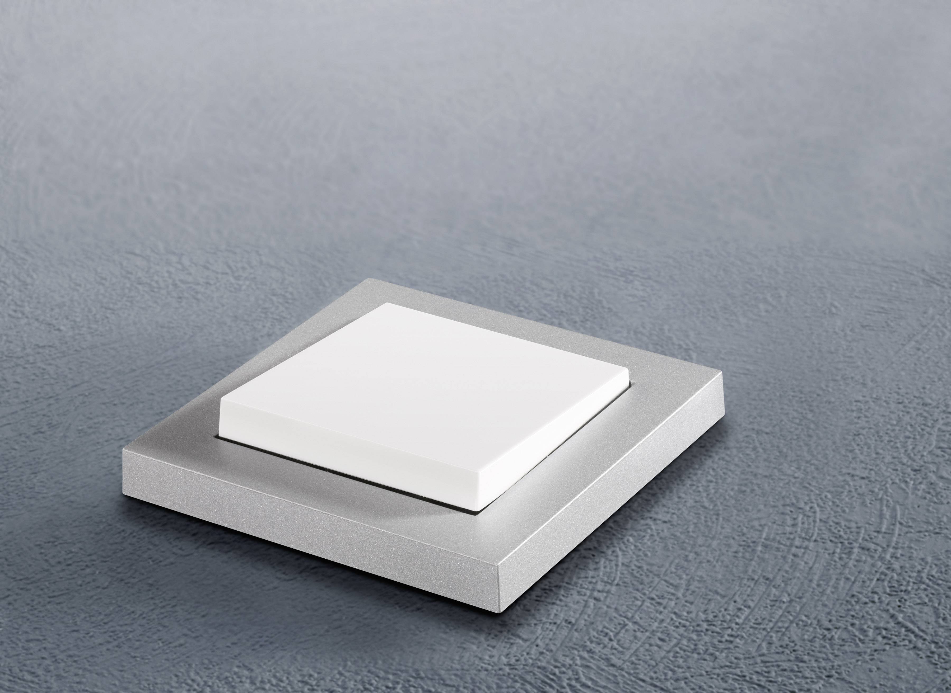 Čtvercový stříbrný vypínač s plochou bílou klávesou na strukturované šedé stěně.