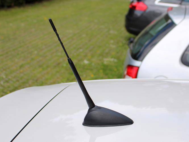 Autoanténa na bílé střeše vozidla, zaparkovaného na travnaté ploše, vedle další automobil.