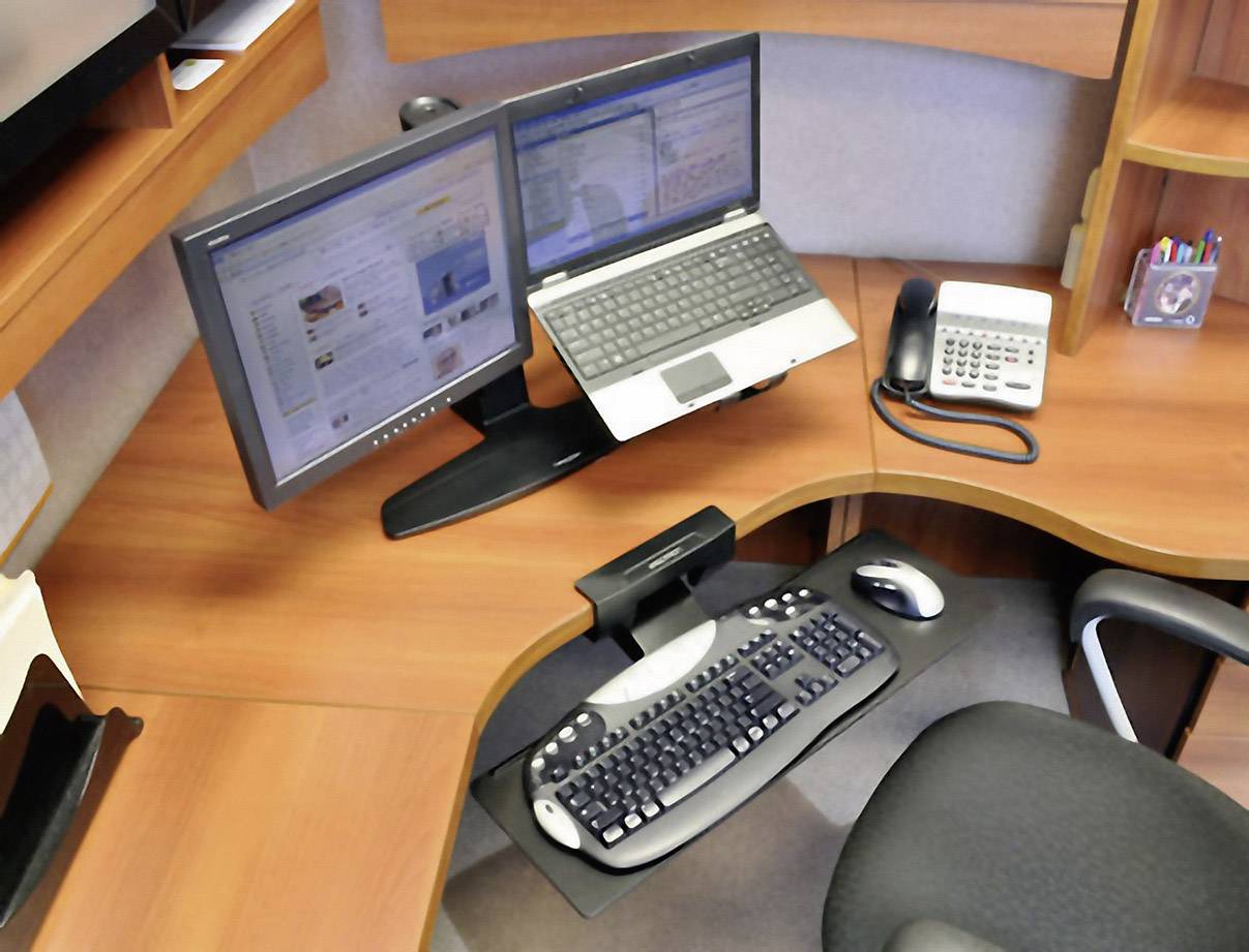 Moderní kancelářský stůl se dvěma monitory, laptopem, telefonem, klávesnicí a myší. Pracoviště působí organizovaně a funkčně.