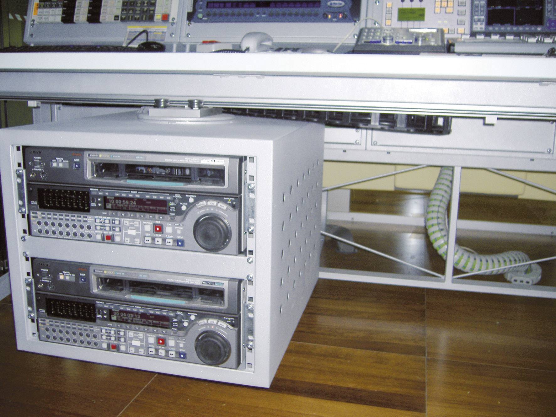 Nahrávací studio s dvěma naskládanými audiopřehrávači na dřevěné podlaze pod psacím stolem. V pozadí jsou viditelné kabely.