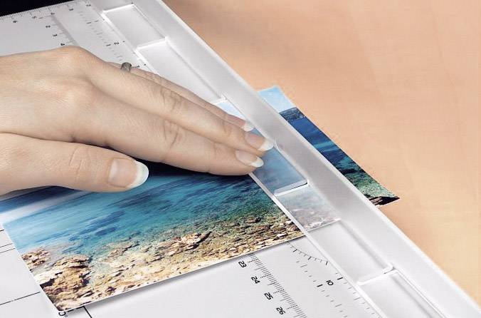 Jedna ruka řeže fotografii pobřežní krajiny pomocí řezáku. Pravítko ukazuje centimetry a palce.