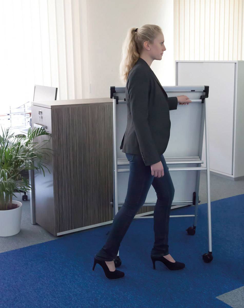 Žena v elegantním oblečení posunuje bílou tabuli na kolečkách kancelářským prostorem s modrým kobercem a rostlinami v pozadí.
