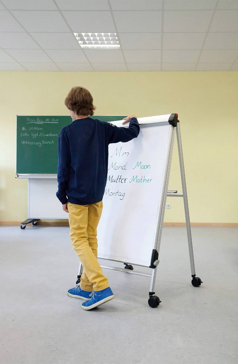 Chlapec stojí před bílou tabulí s písmenem 'M'. V pozadí je tabule s nápisem a křídovými kresbami.