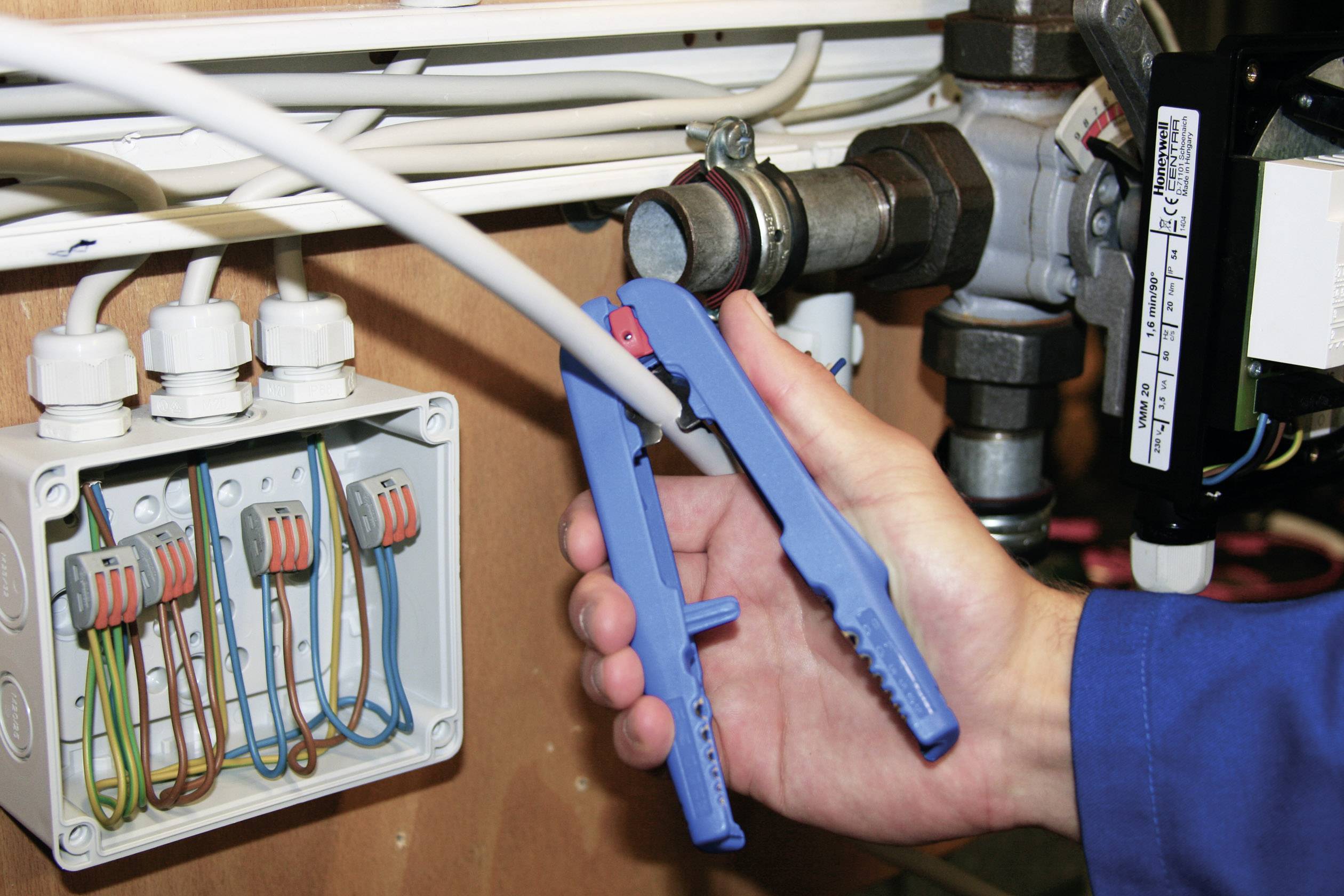 Osoba drží modré nářadí, aby upevnila kabel v elektrickém rozvodném boxu. V pozadí jsou viditelné trubky a ventily.