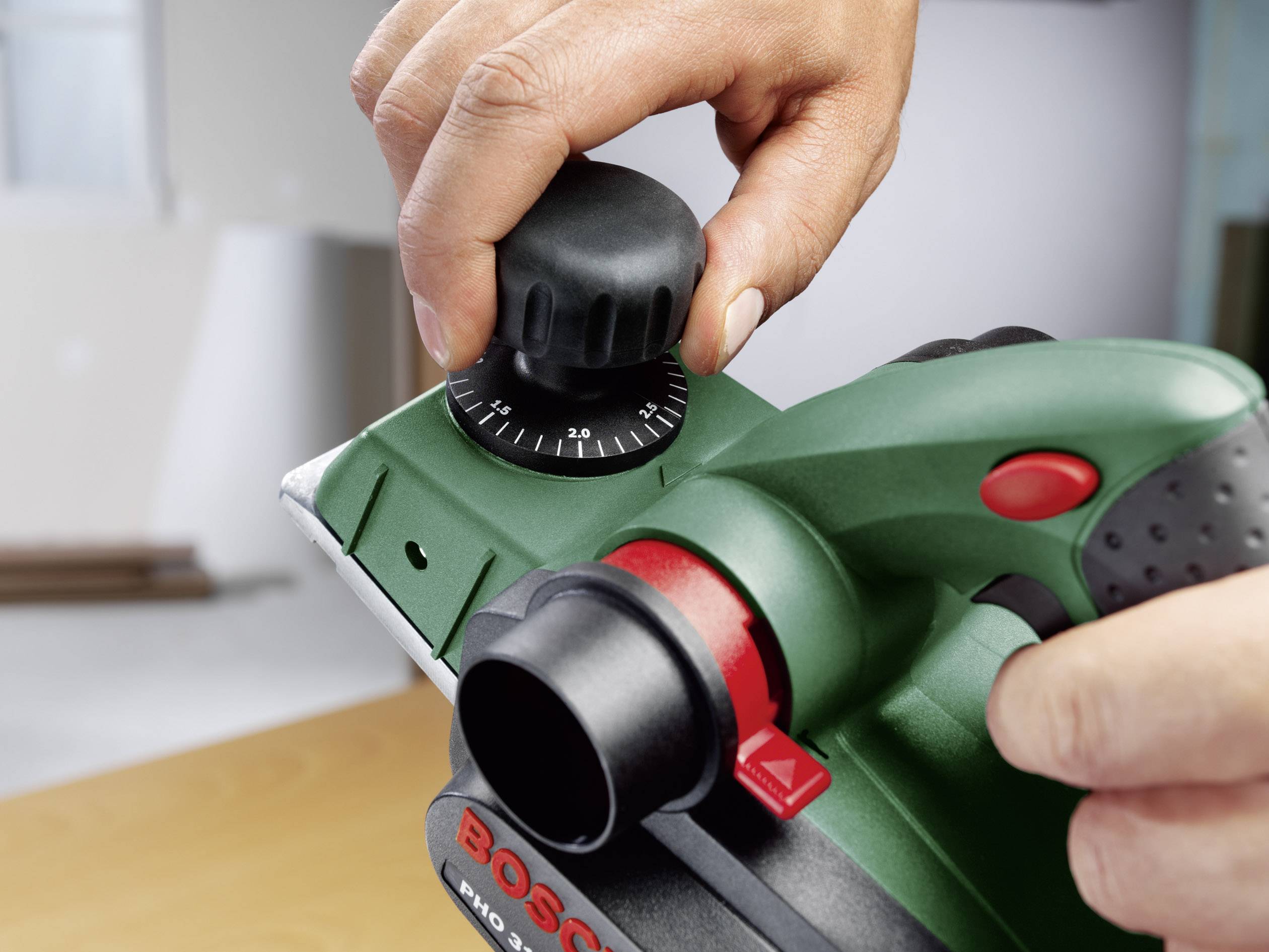 Osoba nastavuje zelený elektrický hoblík otáčením nastavovacího knoflíku. Zařízení je umístěno na desce stolu.