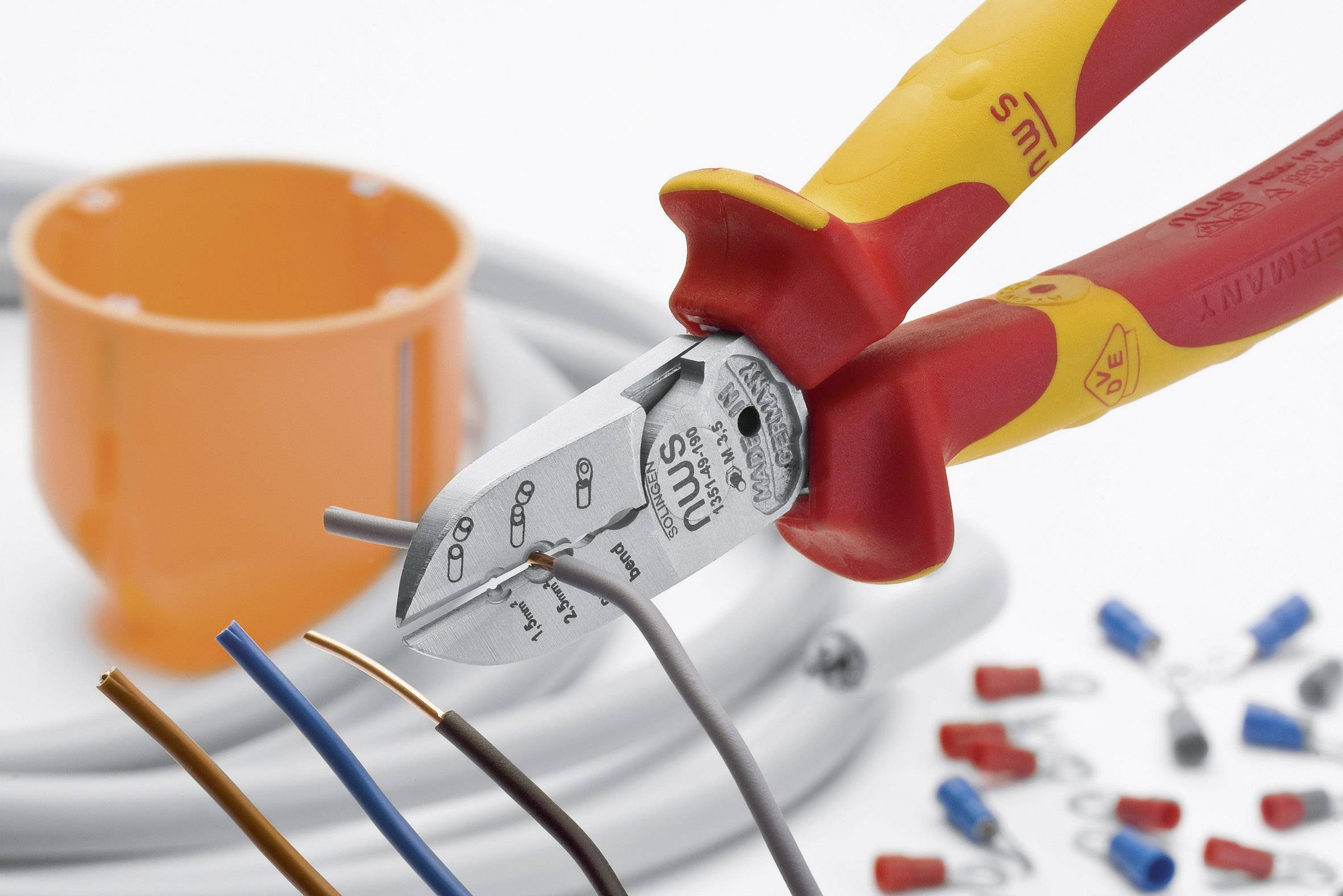 Zange řeže modrý kabel. V pozadí kabely, oranžová rozvodná krabice a barevné kabelové konektory. Symbolizuje elektroinstalační práce.