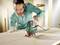 Muž s ochrannými brýlemi a rukavicemi obsluhuje zelenou přímočarou pilu, která řeže na dřevěné desce.