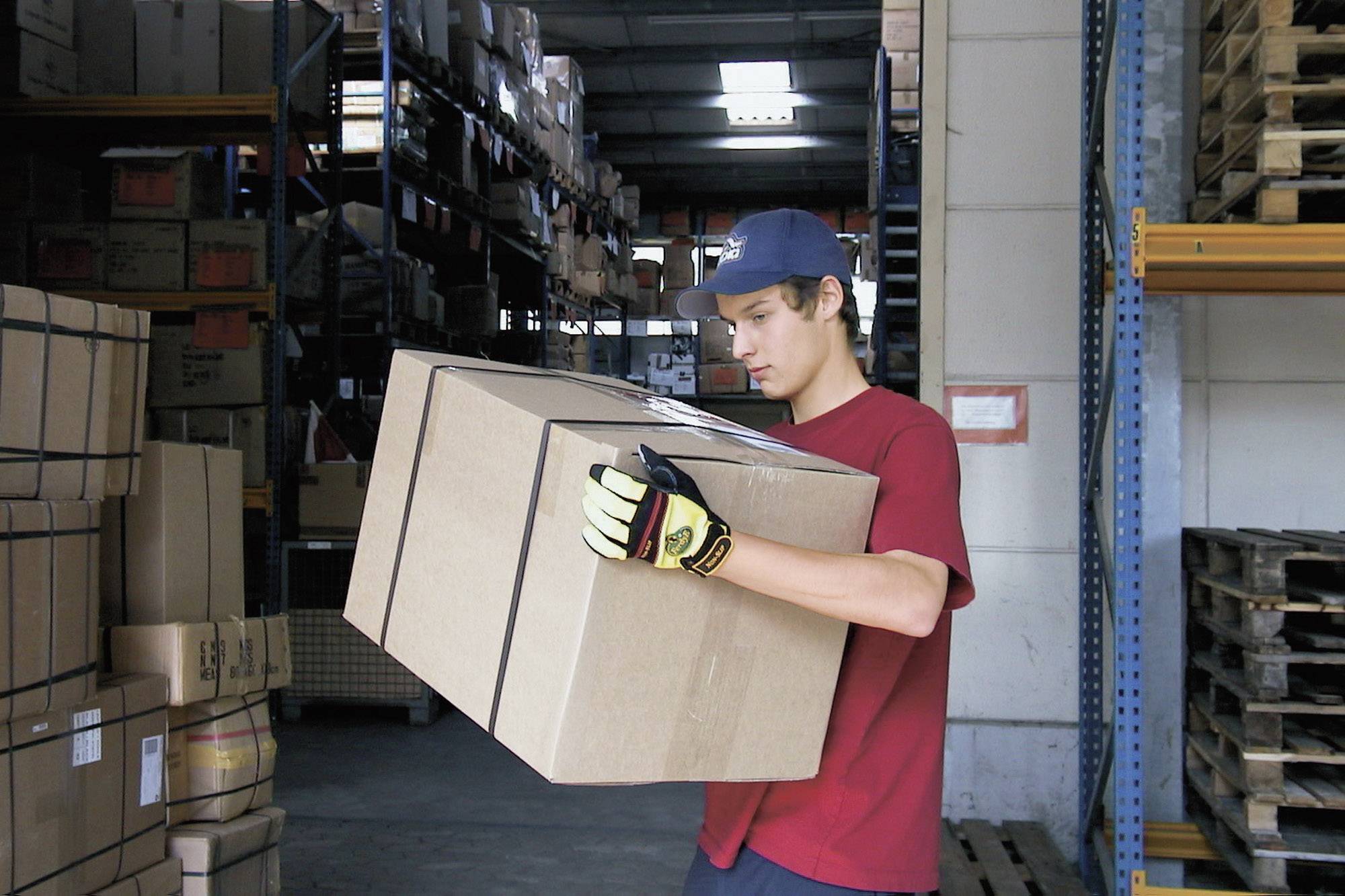 Muž v červeném tričku a modrém klobouku nese velkou hnědou zásilku ve skladu s mnoha regály v pozadí.