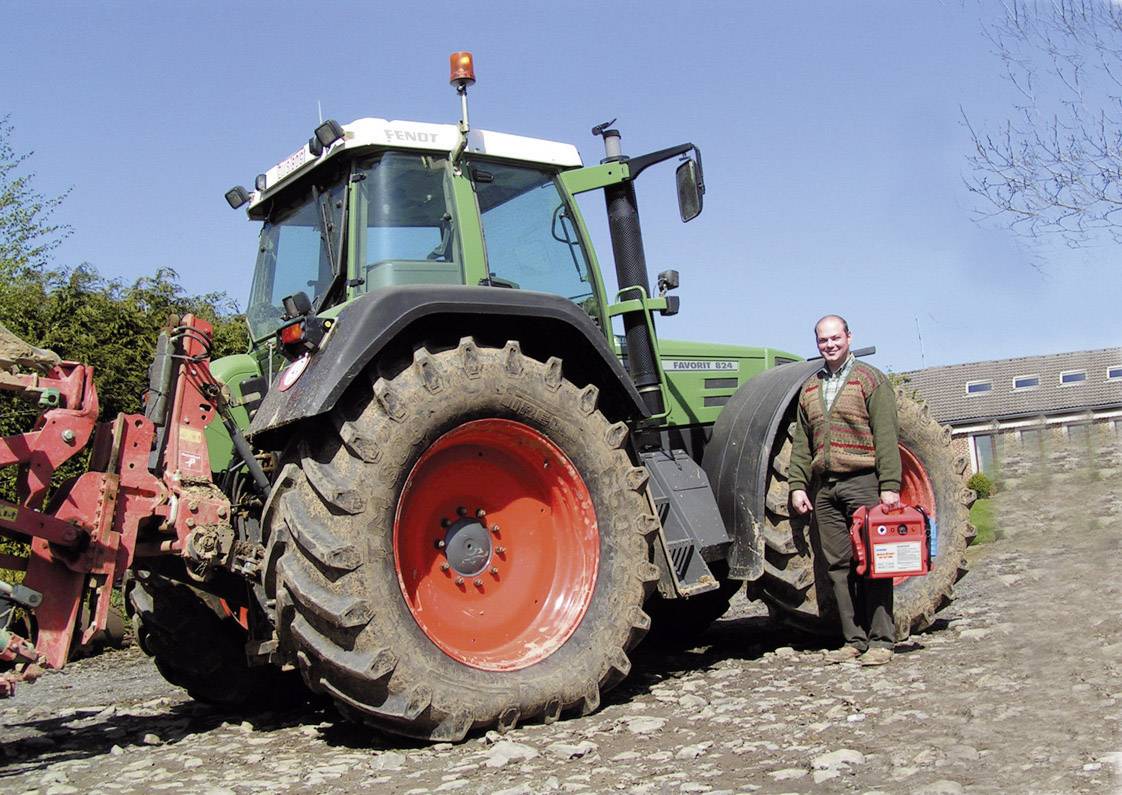 Muž stojí vedle velkého traktoru na dvoře. Traktor je zelený s velkými koly v červeno-oranžové barvě. Dvůr je kamenitý.