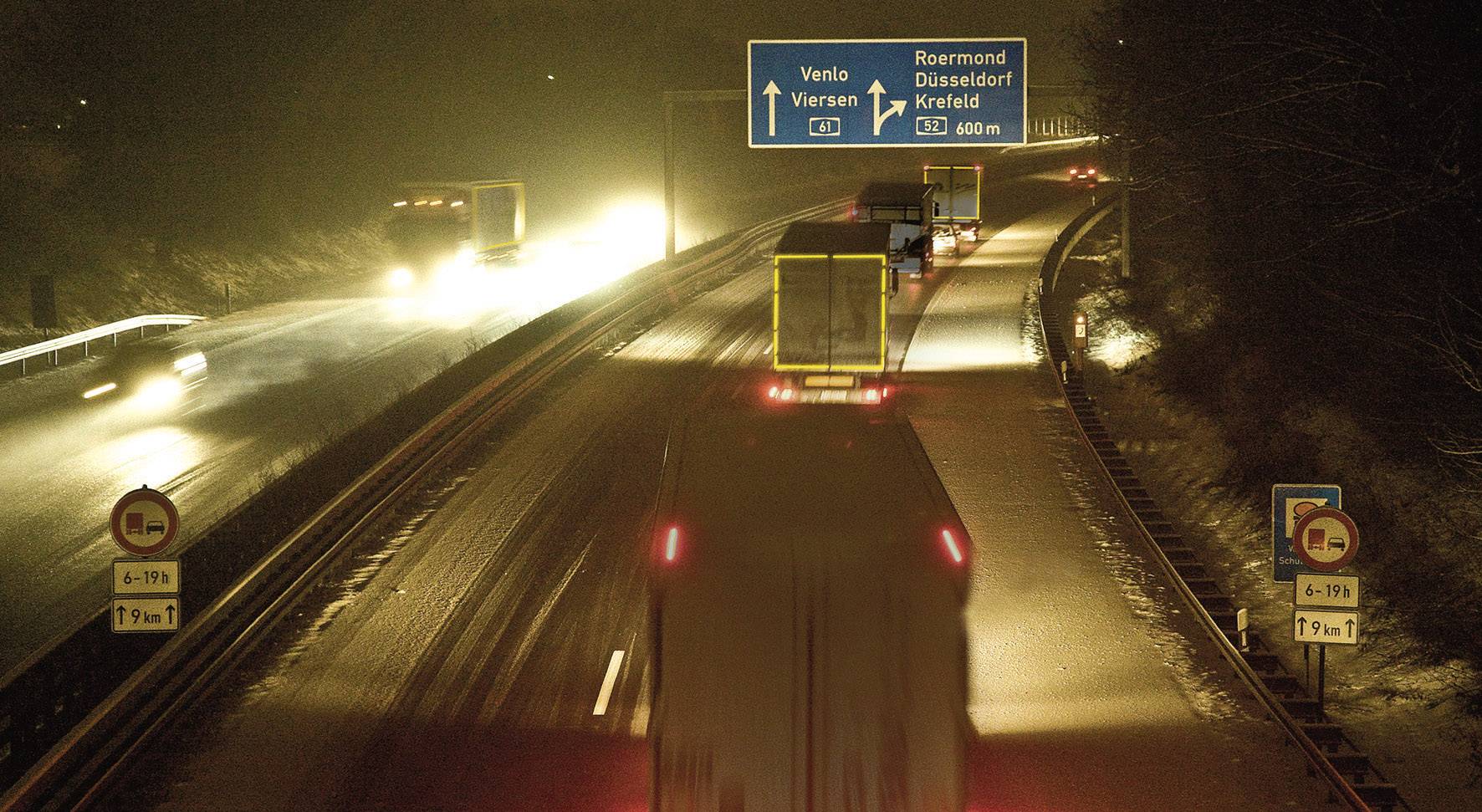 Nákladní automobily jedou v noci po dálnici. Dopravní značka ukazuje směry na Venlo, Düsseldorf a Krefeld.