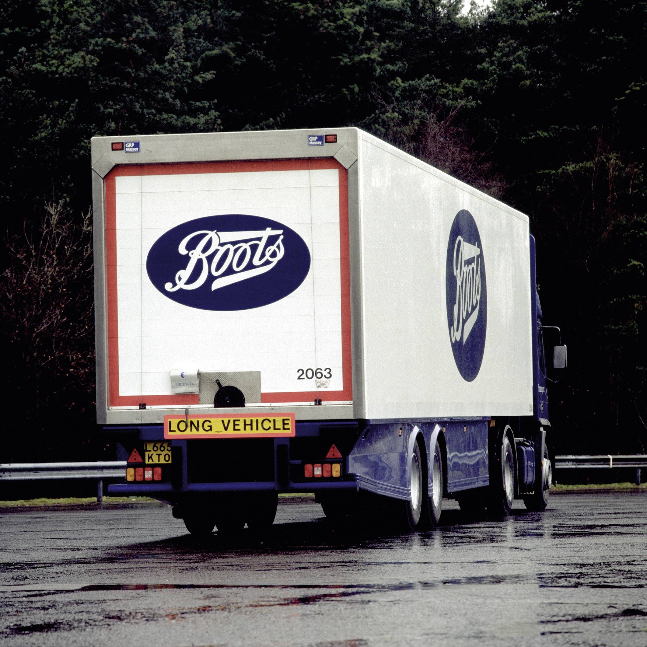 Nákladní automobil s nápisem 'Boots' na parkovišti obklopeném stromy.