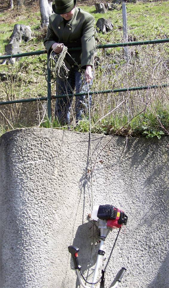 Osoba vytahuje motorovou sekačku pomocí lana na vysokou zeď. V pozadí je tráva a plot.