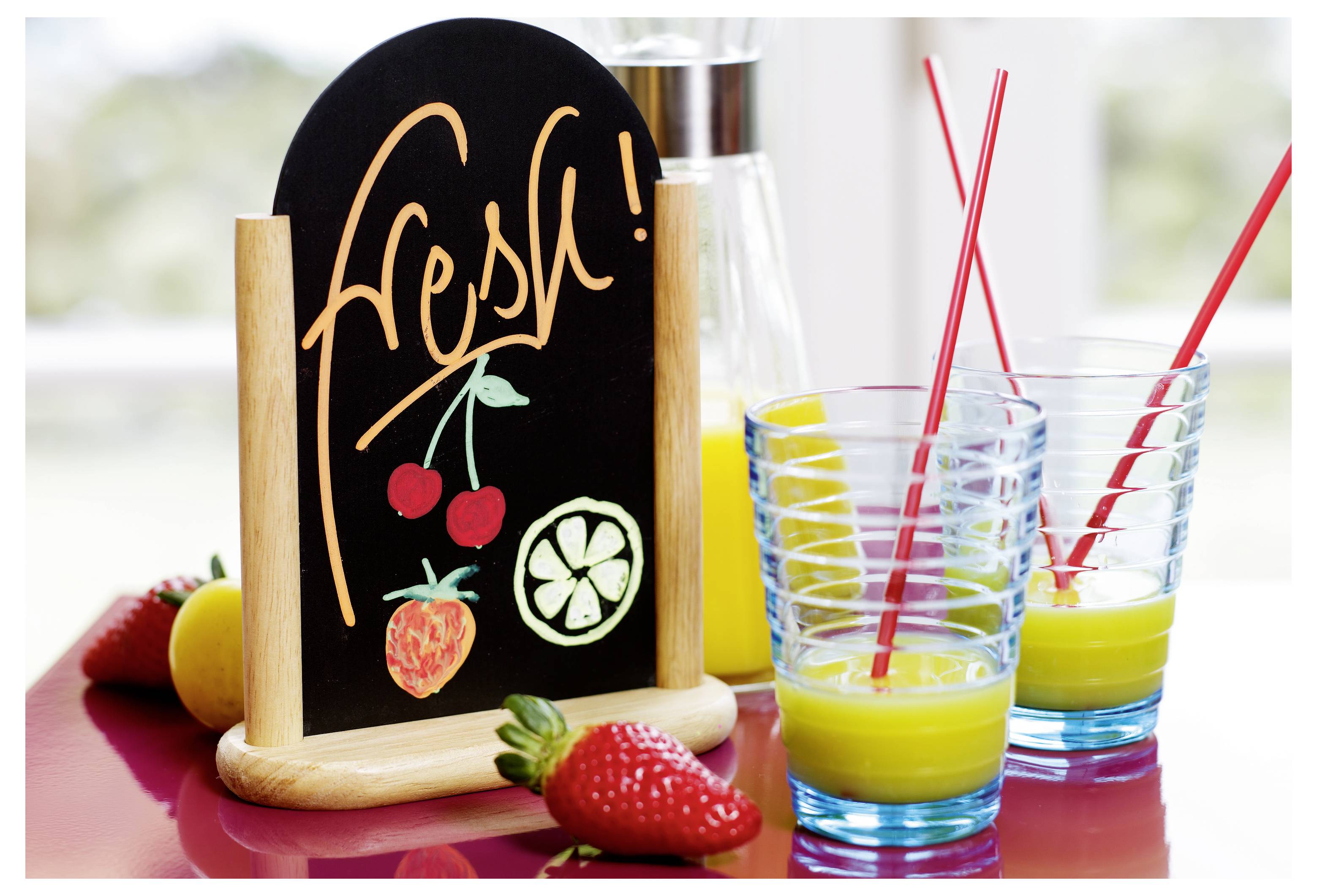 Frisk juiceudstilling med en tavle med teksten 'frisk', jordbær, en citron, skiver af lime og to glas gul juice med røde sugerør.