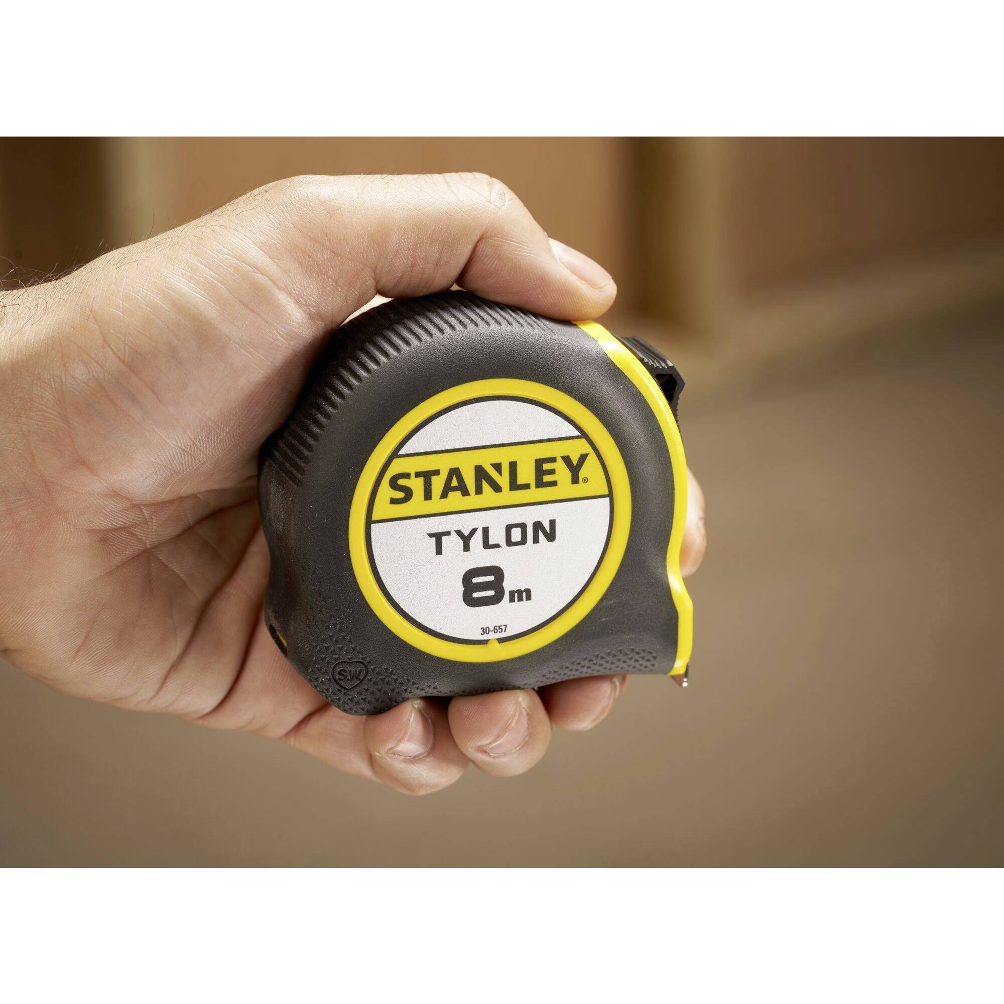 En hånd holder et målebånd fra Stanley, som kan måle op til 8 meter. Hylsteret er sort med gule detaljer.