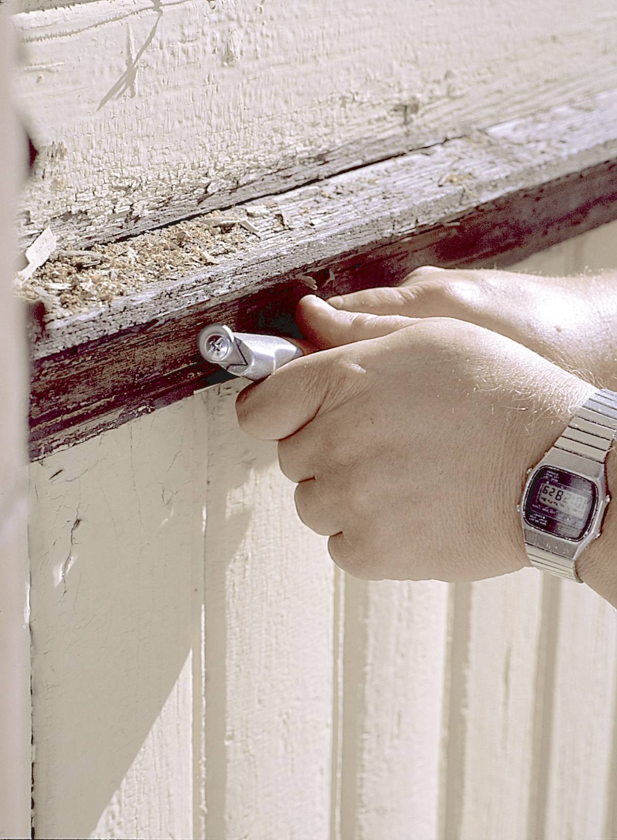En person arbejder med en skruetrækker på en trævinuesramme, bærer et armbåndsur, og en lysebrun træpanel er synlig.