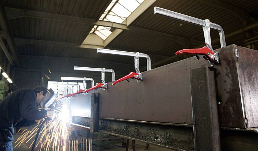 En arbejder svejser et stort metalstykke i en værkstedhal, fastgjort med flere røde klemmer. Gnister sprøjter.