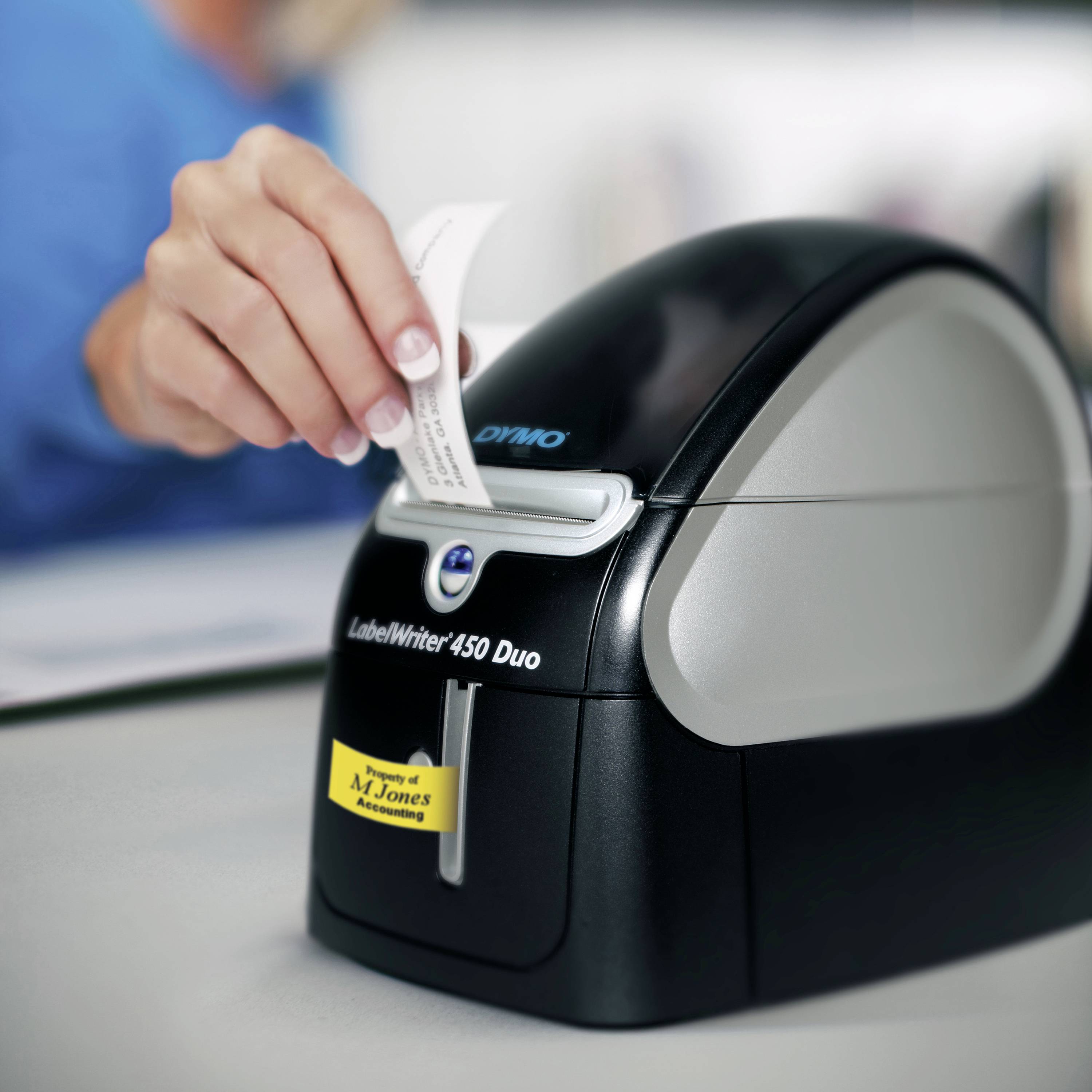 En person betjener en etiketprinter, der udskriver en etiket. På bordet står printeren ved navn 'LabelWriter 450 Duo'.