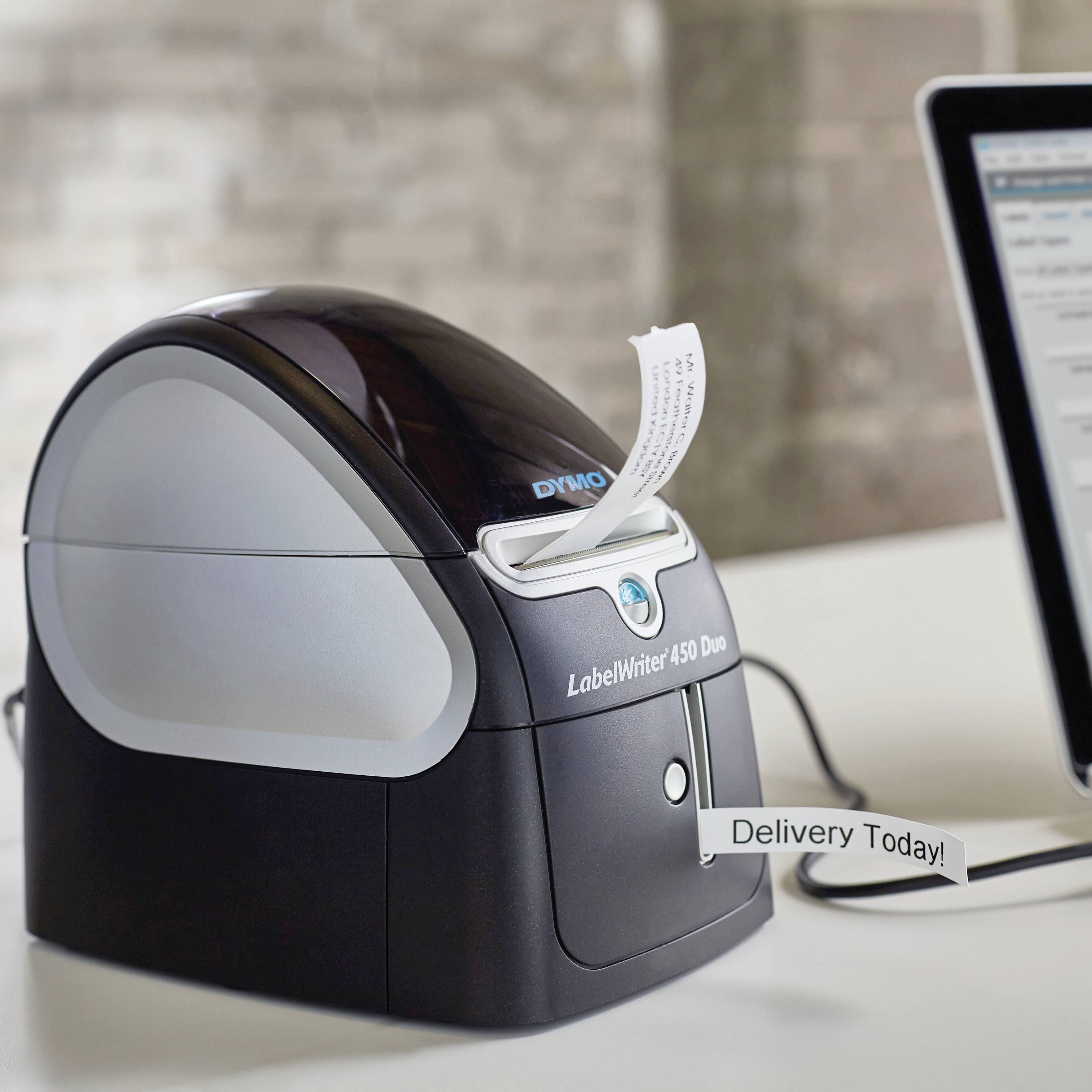 En labelprinter udskriver et mærkat med teksten 'Leveres i dag'. En computerskærm er synlig i baggrunden.