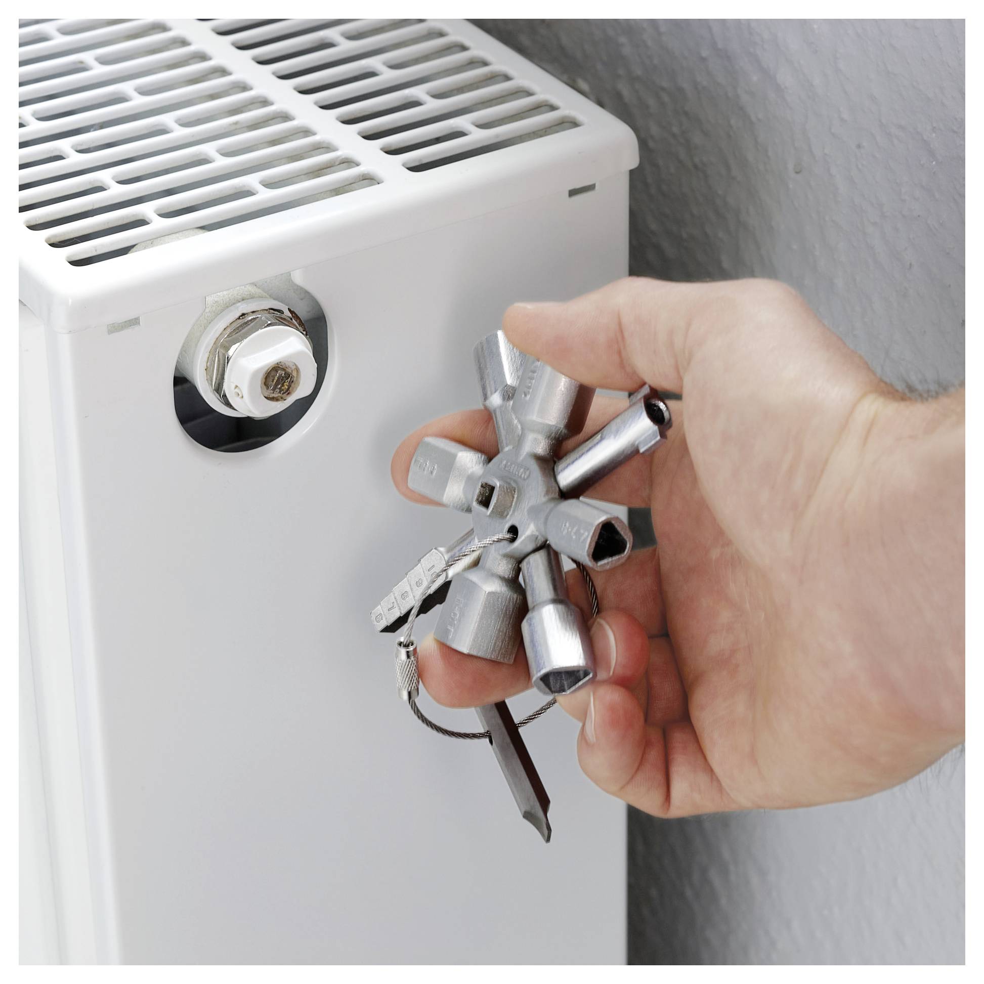 En hånd, der bruger et multiværktøj til at lufte en radiator, og justerer ventilen på en hvid radiator med en væg i baggrunden.