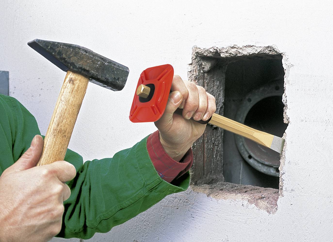 Person i grøn jakke bruger hammer og mejsel til at bearbejde et hul i væggen. Sten eller beton fjernes.