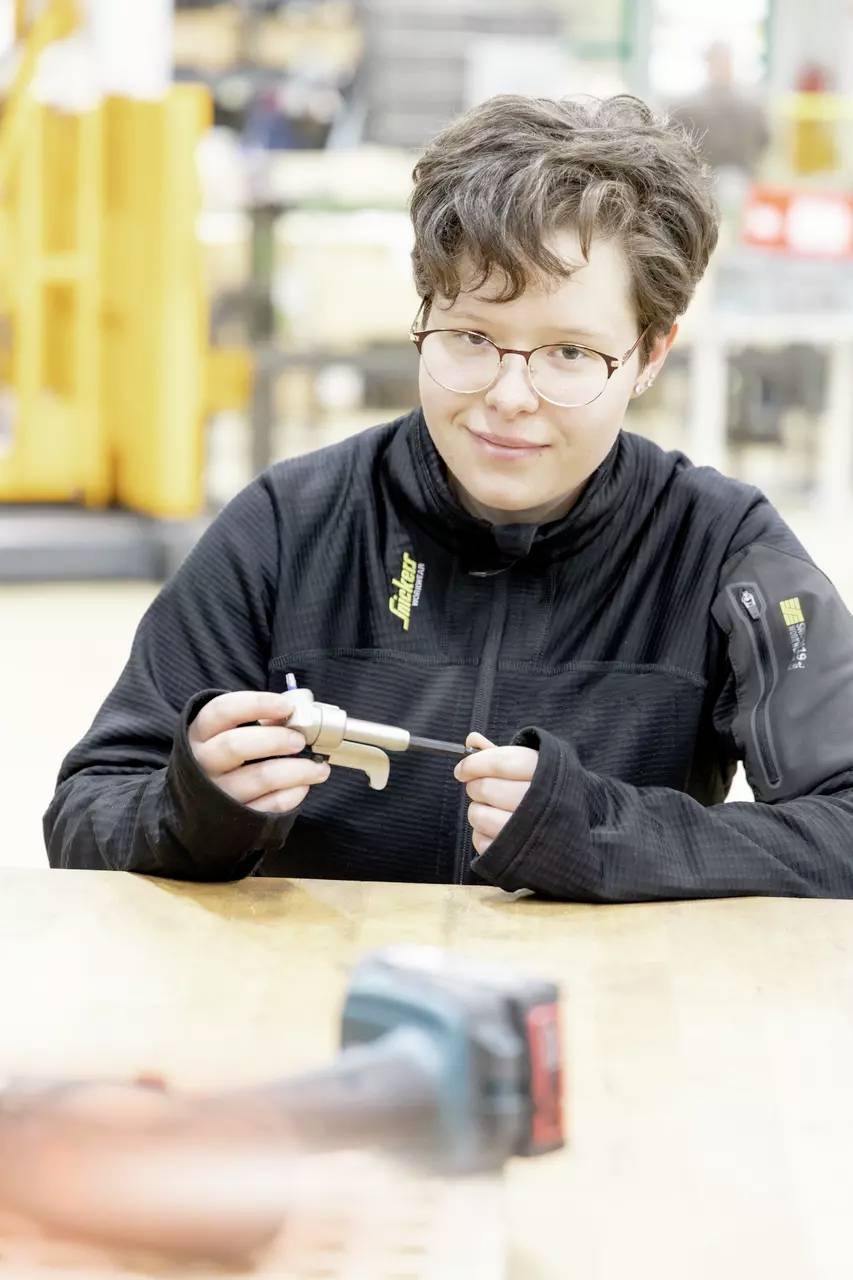 En person med kort hår og briller sidder ved et bord og holder et lille metalværktøj og en bolt, iført en sort jakke.