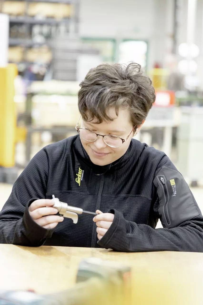 En person med kort hår og briller undersøger en metalgenstand ved en arbejdsplads og fokuserer intenst på objektet. Hylder og udstyr er slørede i baggrunden.
