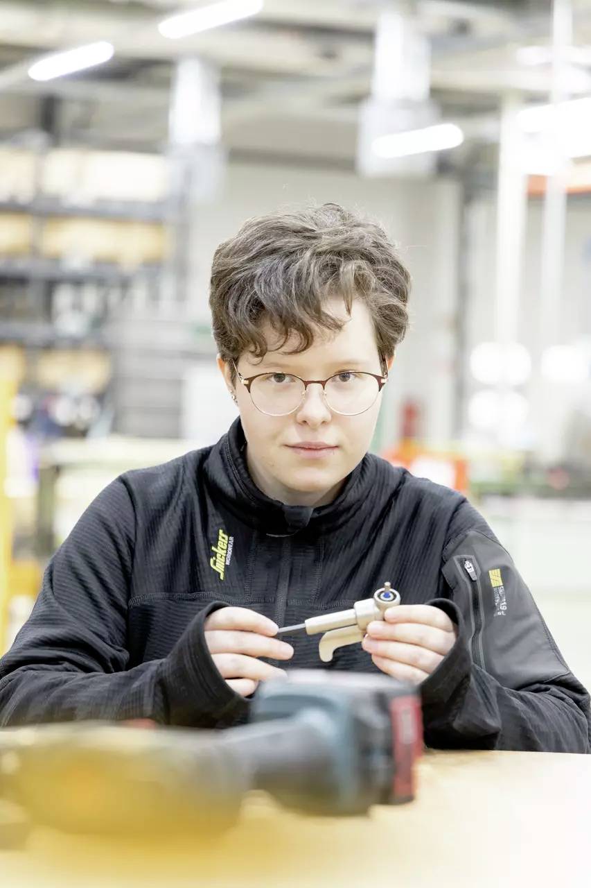 En person iført briller og en sort jakke undersøger en lille mekanisk del i et industrielt værkstedsmiljø.