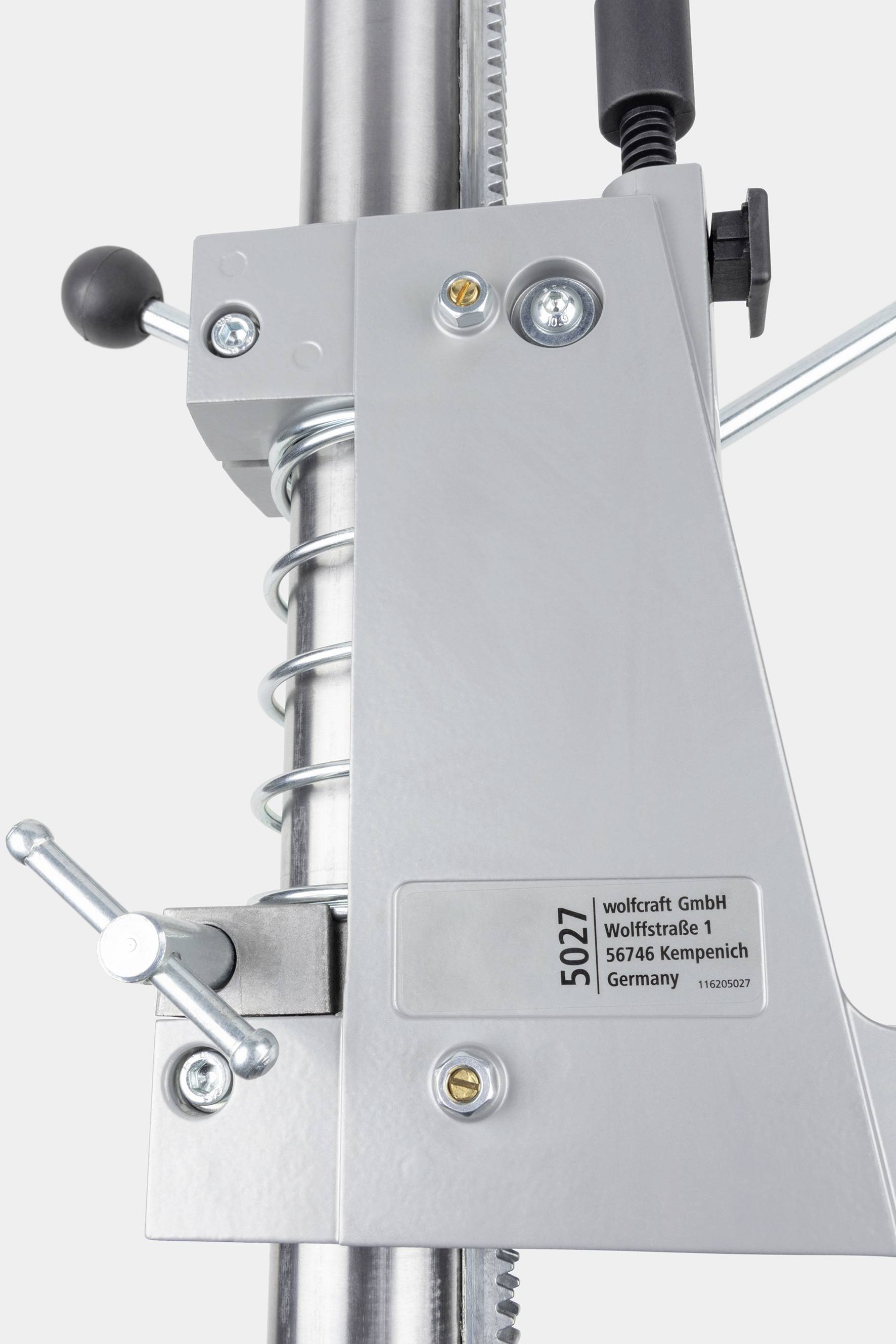 Maskinkomponent med metalarm og fjeder, mærket med '5027' og adressen 'wolfcraft GmbH, Kempenich, Tyskland'.