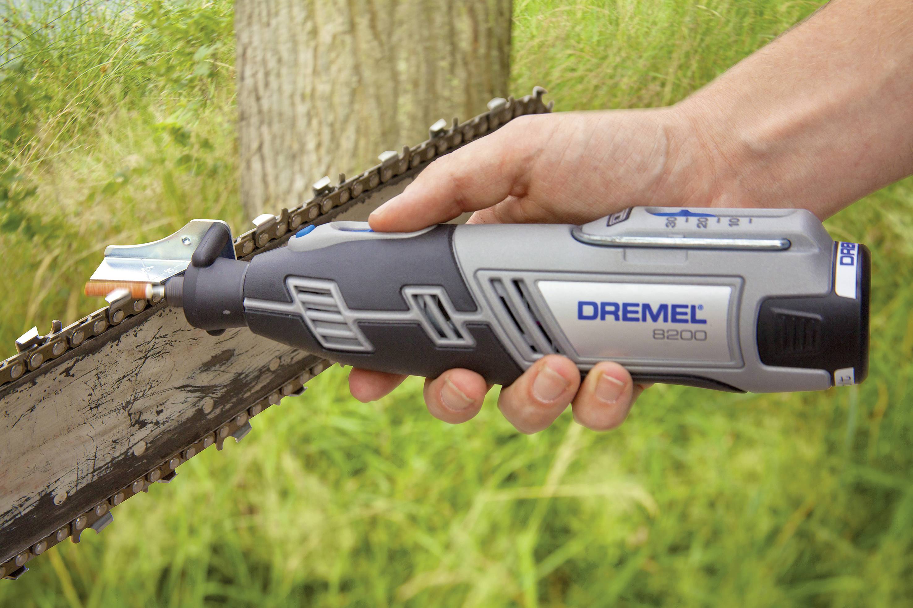 En person holder en Dremel 8200-værktøj op mod en kædesavskæde i en grøn omgivelse.
