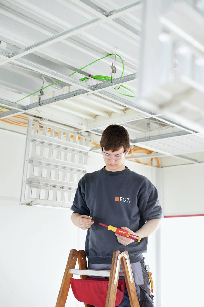 En elektriker står på en stige og arbejder med loftsinstallationen i et industrielt rum.