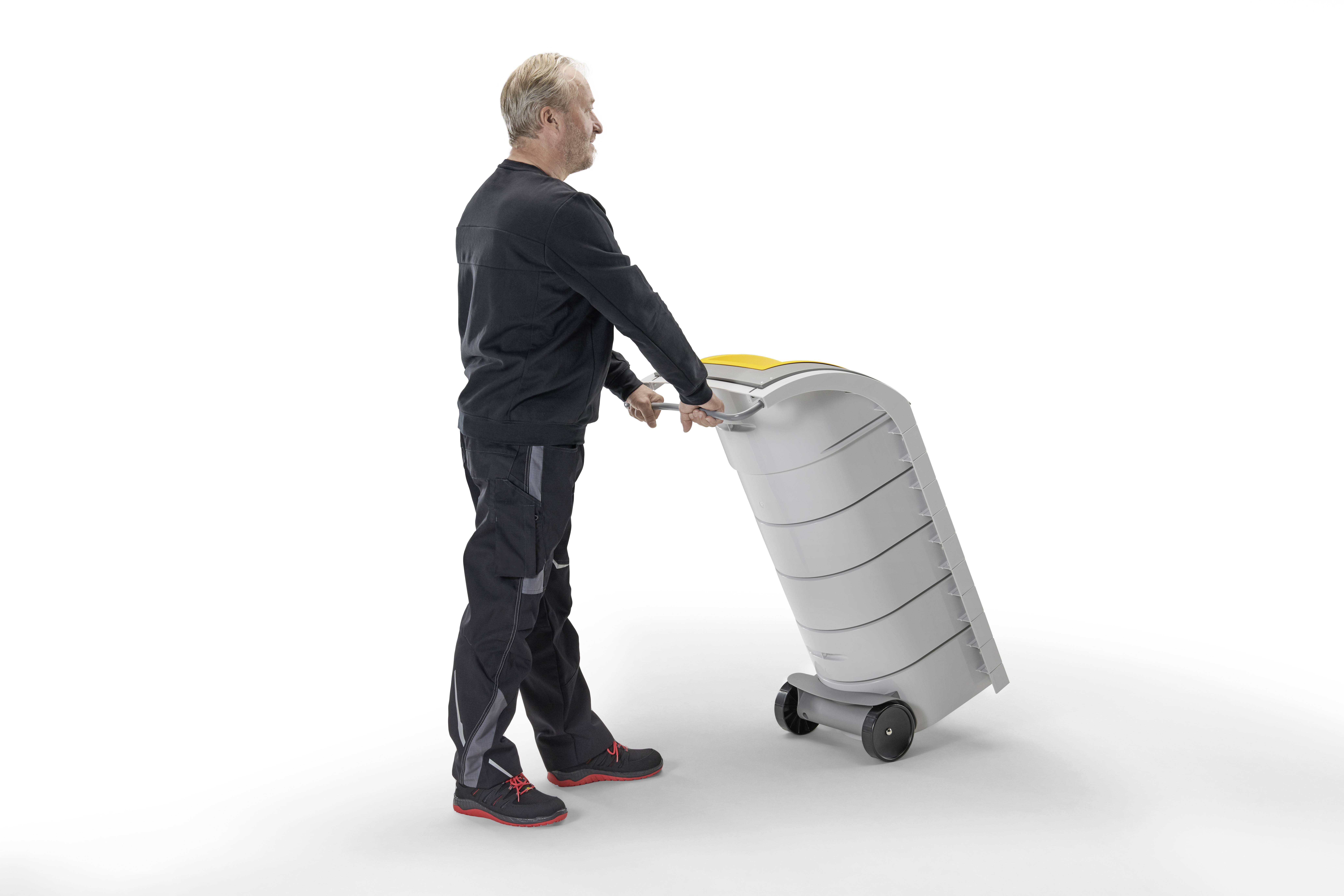 En mand skubber en grå affaldscontainer med hjul. Han bærer sorte klæder og har gråt hår.