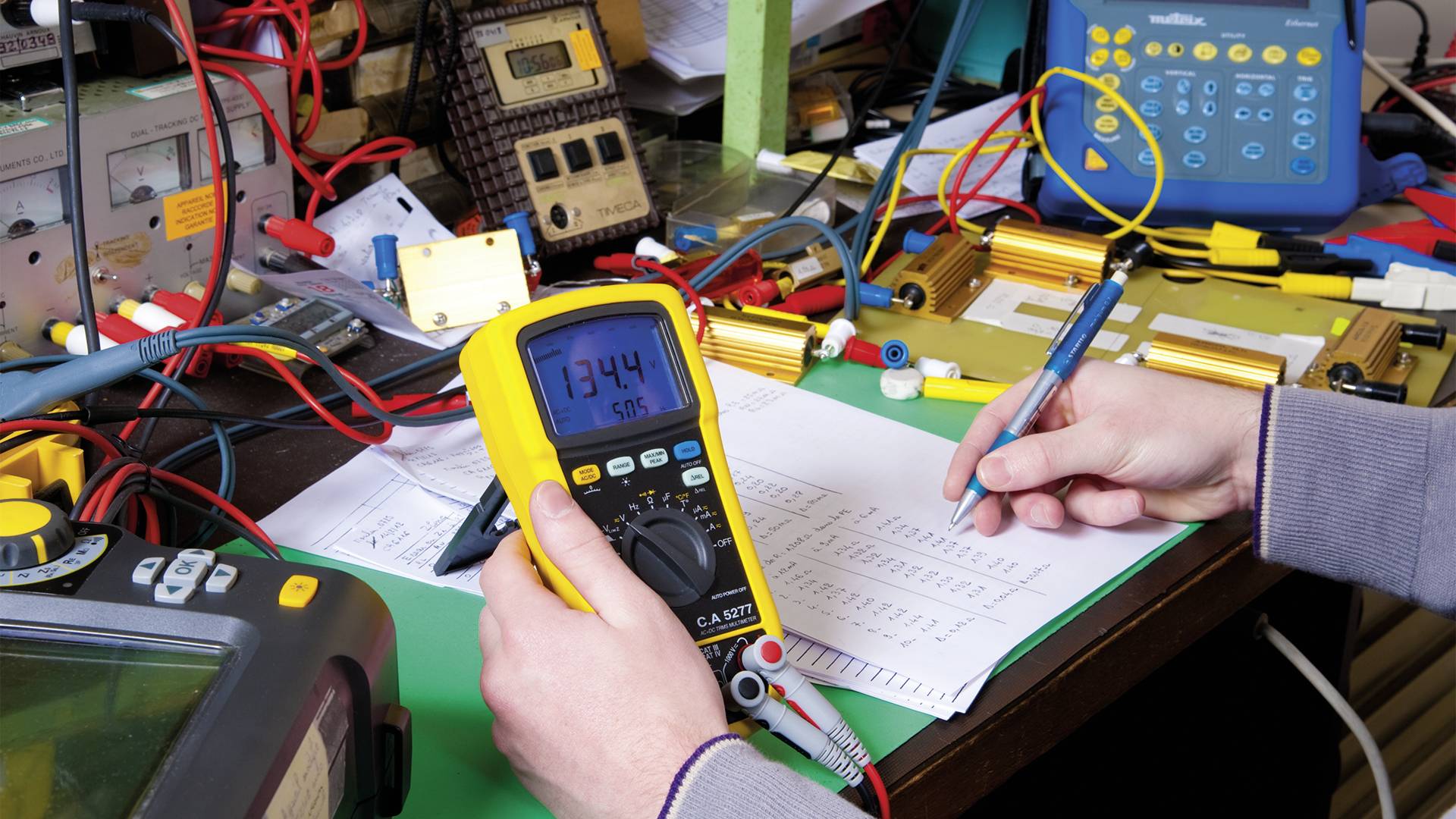 En person måler spænding med et multimeter på en elektronisk enhed, mens vedkommende tager noter. Et bord fyldt med kabler og udstyr.