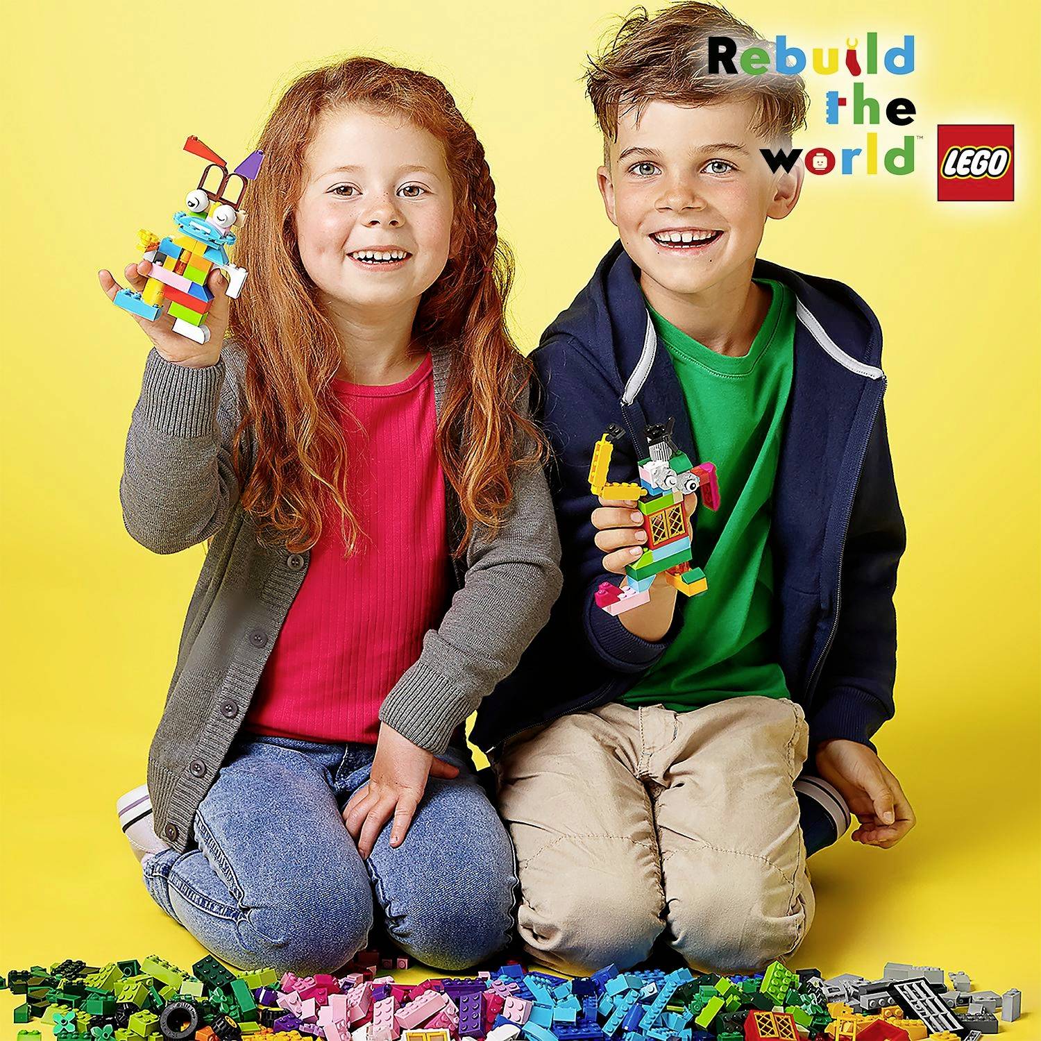To glade børn sidder foran en bunke farverige LEGO-klodser. Begge holder LEGO-figurer. Øverst til højre står der 'Rebuild the world'.