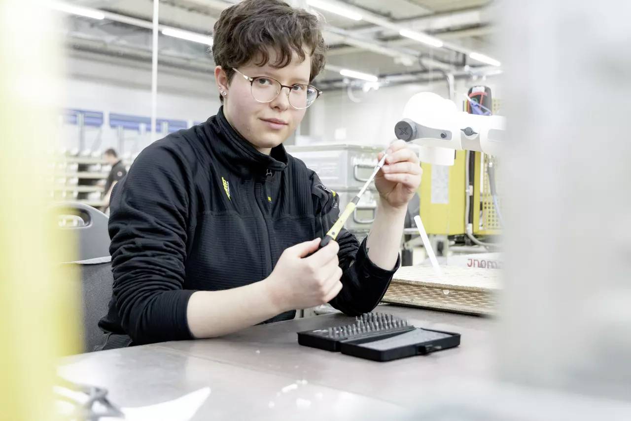 En person arbejder fokuseret i et fabriksmiljø og holder en elektronisk komponent. I baggrunden ses maskiner.