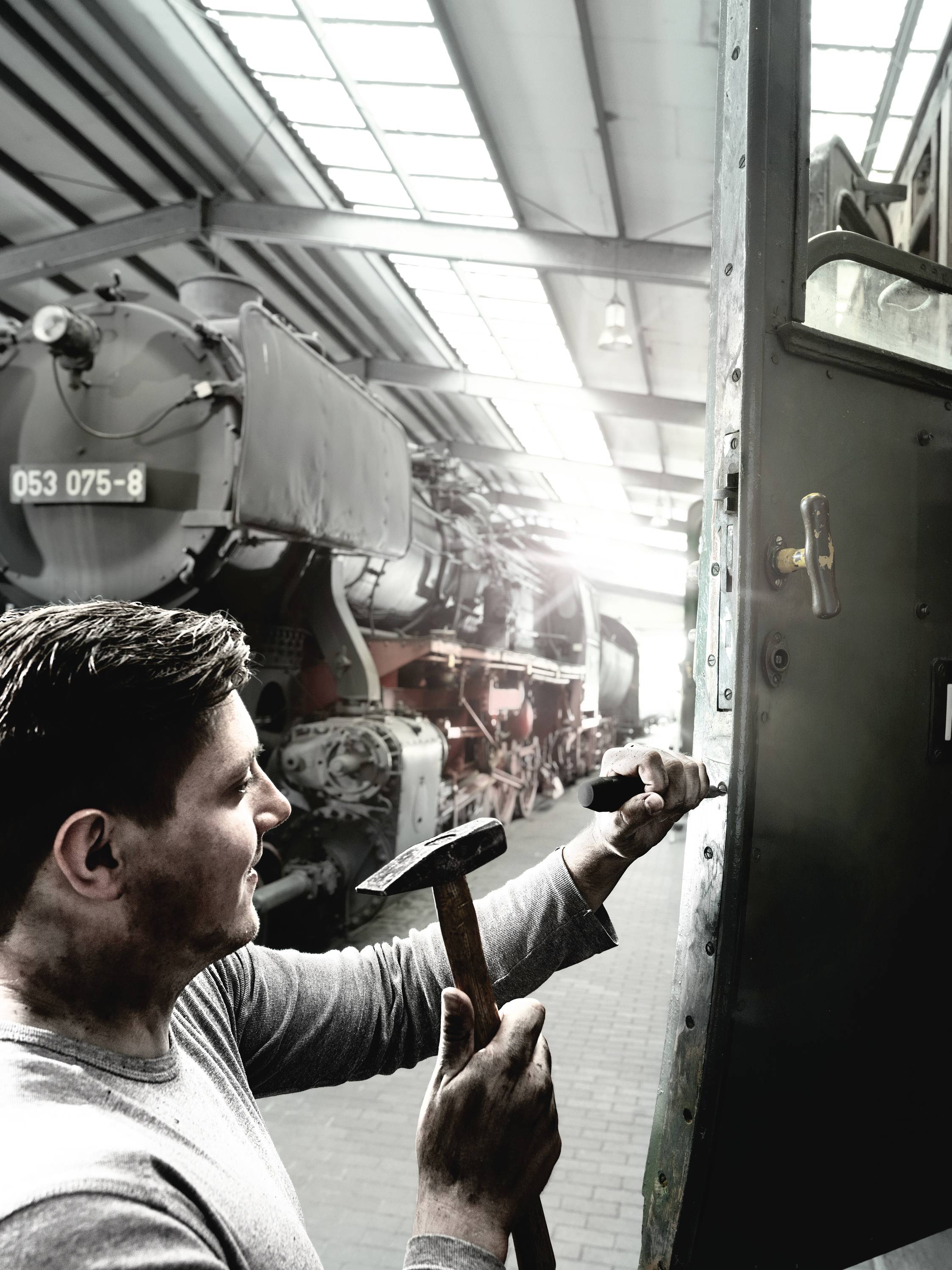 En mand med en hammer arbejder på døren til et gammelt lokomotiv i en hal fuld af historiske tog.