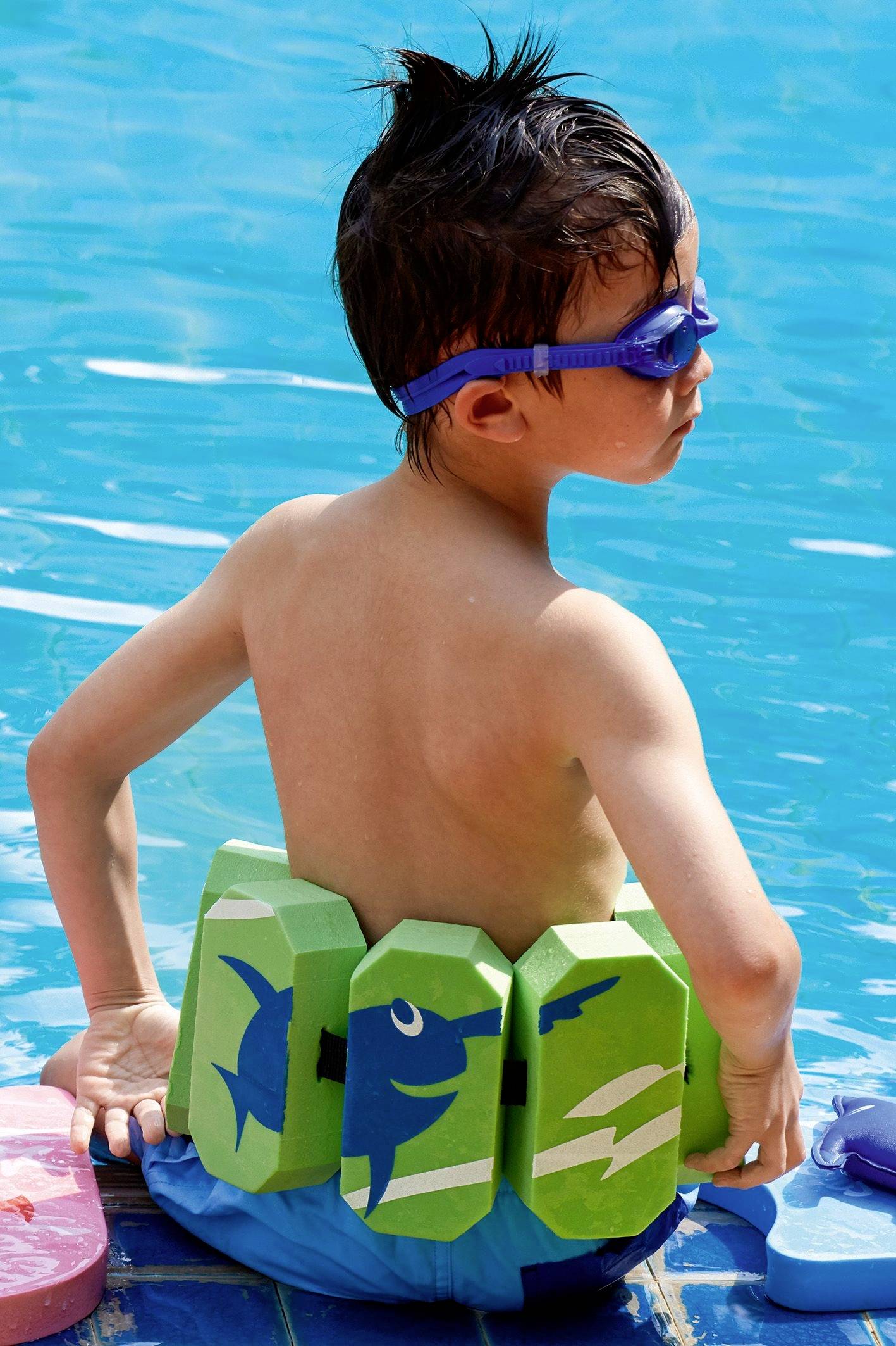 Et barn med svømmebriller sidder ved kanten af swimmingpoolen, iført en grøn svømmevest med haj-motiv og er klar til at svømme.