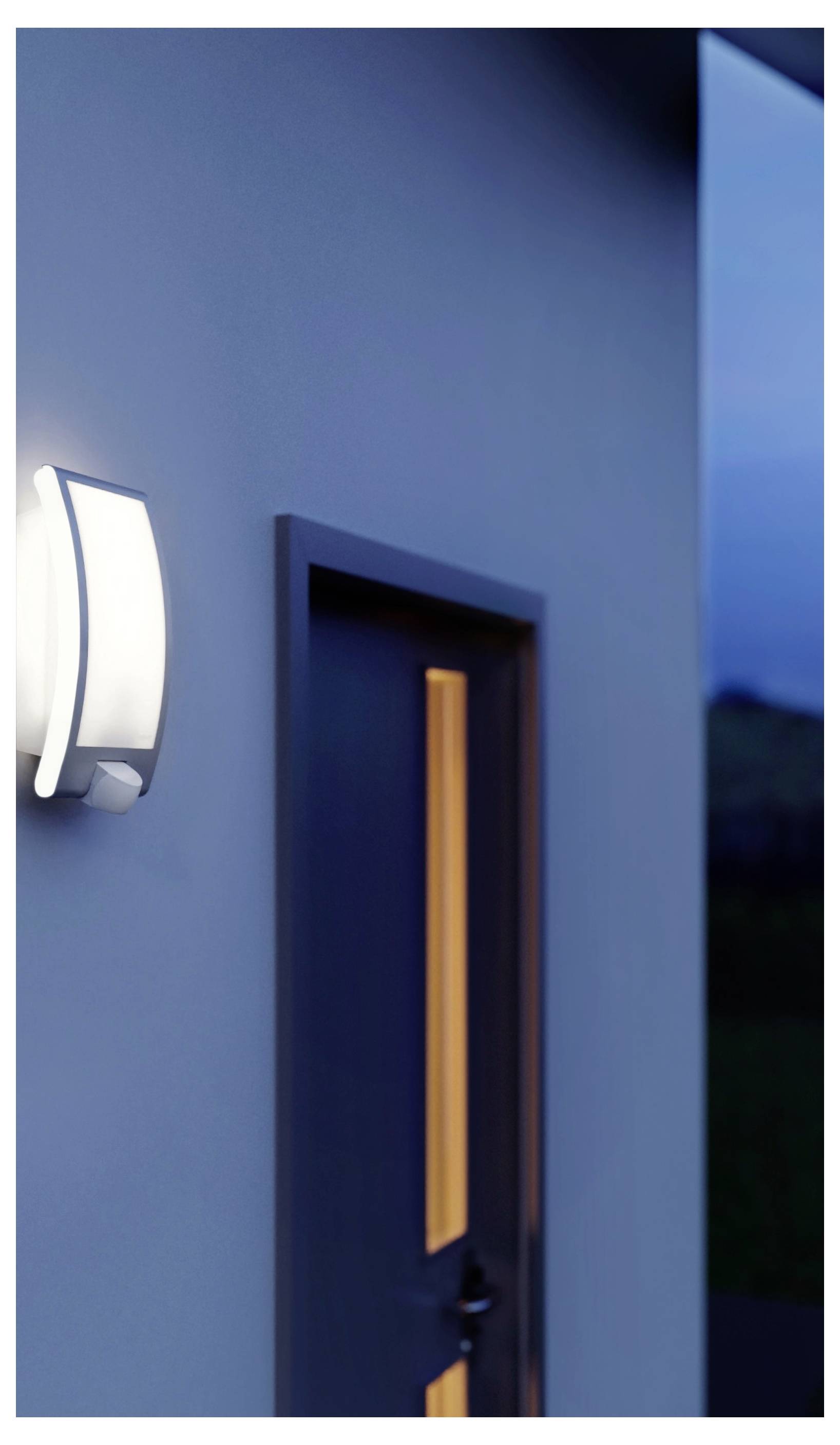 Ydervæg af en moderne bygning med en oplyst væglampe og en delvist åben dør i skumringen