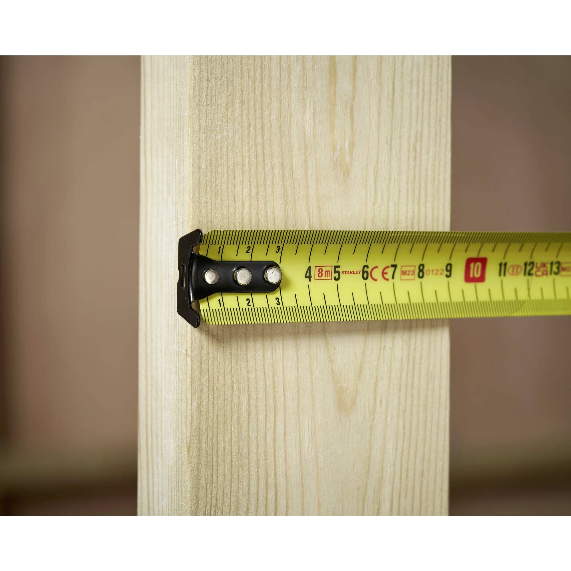 Et gult målebånd måler bredden af et lodret stykke træ. Båndet viser omkring 6 centimeter.