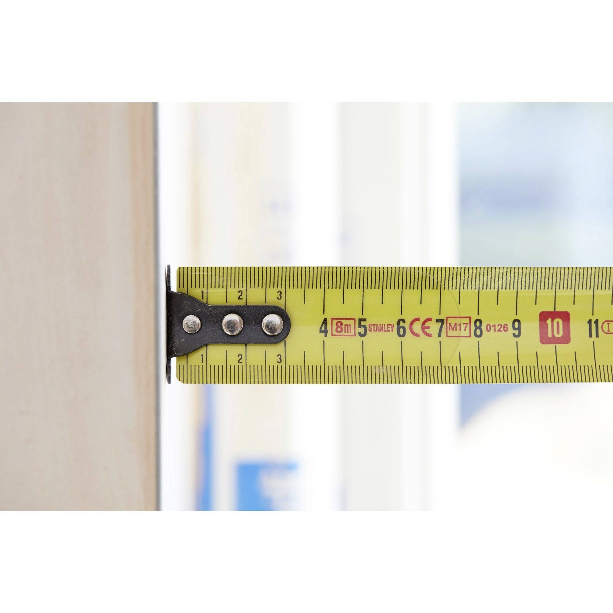 Et målebånd måler bredden af et stykke træ. Den målte bredde er 7,8 cm. Uskarpe detaljer i baggrunden.