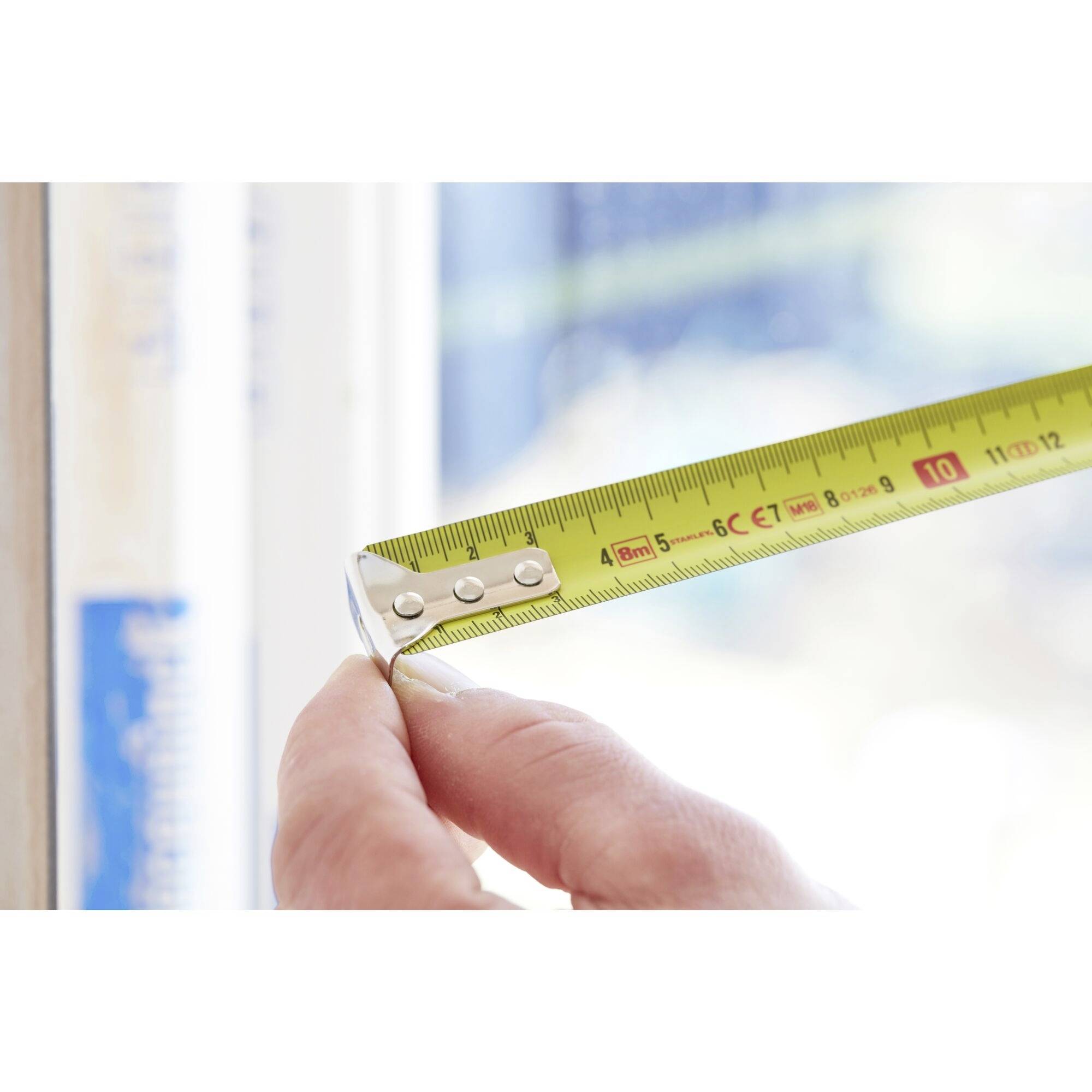 En hånd holder et målebånd, der er placeret langs en vinduesramme. Skalaen viser en måling i centimeter.